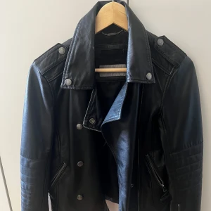 Svart skinnjacka från Calvin Klein Jeans - Snygg svart skinnjacka från Calvin Klein Jeans i storlek XS. Jackan har en klassisk bikerstil med dragkedja och knappar, samt detaljerade sömmar på axlarna. Perfekt för en cool och stilren look.