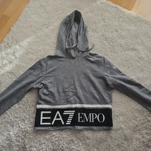 Grå hoodie från Emporio Armani - Säljer en snygg grå hoodie från Emporio Armani med EA7-logga. Tröjan har långa ärmar och en huva. Den har en bred svart rand med vit text längst ner. Perfekt för en avslappnad stil eller snyggt till jeans. 