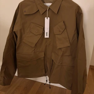 Brun jacka från Uniform Bridge - Säljer en snygg brun jacka från Uniform Bridge med klassisk design. Jackan har långa ärmar och flera praktiska fickor med knappar framtill. Perfekt för en stilren look.                          Butikspris ca 4000 kr. Helt ny med tags storlek L