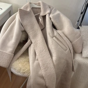 Beige kappa med teddyfoder - Säljer en stilren beige kappa med mjukt teddyfoder. Kappan har en klassisk krage och ett bälte i midjan för en snygg passform. Perfekt för kyliga dagar!