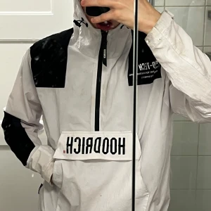 Vit och svart vindjacka från Hoodrich - Säljer en stilren vit och svart vindjacka från Hoodrich med dragkedja och huva. Jackan har en stor ficka framtill med Hoodrich-logga och svarta detaljer på ärmar och axlar. Perfekt för en sportig look.