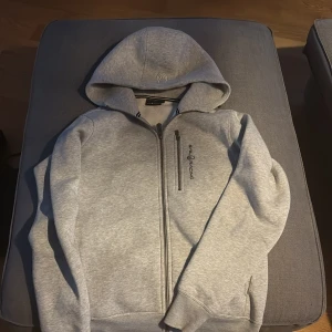 Grå hoodie från Sail Racing - Säljer en stilren grå hoodie från Sail Racing med dragkedja och en praktisk bröstficka. Perfekt för kyliga dagar med sin mjuka insida och justerbara huva. Passar både till vardags och mer avslappnade tillfällen.