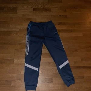 Blå träningsset från McKenzie - Säljer ett blått träningsset från McKenzie med byxor och huvtröja. Byxorna har elastisk midja och vita detaljer på benen. Huvtröjan har dragkedja och vita paneler på framsidan samt en huva. Perfekt för sport och fritid.