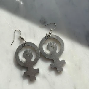 Silverörhängen kvinnosymbol - Snygga silverfärgade örhängen med en feministisk symbol som har en knuten näve i mitten. Perfekta för att göra ett statement och visa ditt stöd för jämställdhet. De är lätta att bära och har en krok för enkel användning.