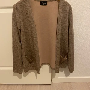 Beige cardigan jacka från Gina - Säljer en stilren beige cardigan jacka från Gina. Jackan har en öppen design utan knappar och två praktiska fickor framtill. Perfekt för lager-på-lager under höst och vår.