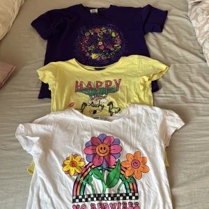 Tre färgglada t-shirts med tryck - Säljer tre t-shirts i olika färger och med glada tryck. En vit t-shirt med blommor och texten 'No Bad Vibes', en gul med texten 'Happy Lucky', och en lila med ett färgglatt mönster. Perfekta för att lysa upp garderoben! 🌼