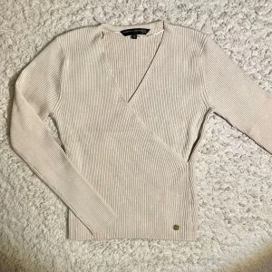 Beige ribbad omlottopp från Pernilla Wahlgren - Snygg beige omlottopp från Pernilla Wahlgren i ribbat material. Toppen har en v-ringad design och långa ärmar, perfekt för en stilren look. Den lilla logotypdetaljen i metall ger en extra touch av elegans.