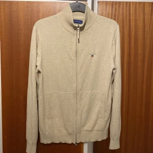 Beige kofta från GANT - Säljer en stilren beige kofta från GANT med dragkedja och broderad logotyp på bröstet. Perfekt för en avslappnad men ändå snygg look. Koftan har långa ärmar och ribbade kanter för extra komfort. Tröjan har används max 5 gånger så den är i helt nytt skick.