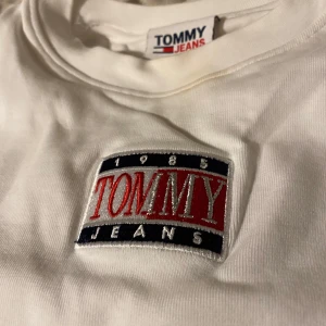 Vit t-shirt från Tommy Jeans - Säljer en stilren vit t-shirt från Tommy Jeans med ett stort, broderat logotypmärke på bröstet. T-shirten har korta ärmar och en klassisk rund halsringning. Perfekt för en casual look!