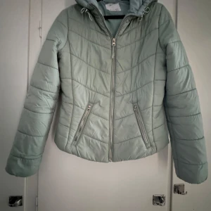 Grön pufferjacka från Bershka - Säljer en snygg grön pufferjacka från Bershka. Jackan har en dragkedja framtill och en praktisk huva. Perfekt för kyligare dagar med sin quiltade design och långa ärmar. Två fickor med dragkedja för säker förvaring.