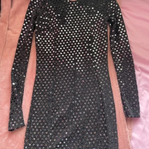 Svart glittrig klänning - Snygg svart glittrig klänning med långärmad design. Perfekt för en kväll ute med vänner. Den har en tight passform som framhäver figuren och ett unikt rutigt mönster som ger en extra touch av glamour.