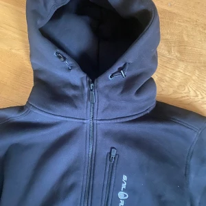 Svart hoodie från Sail Racing - Snygg svart hoodie från Sail Racing med dragkedja och huva.  Hoodien har en bröstficka med dragkedja och justerbar huva för extra komfort. Använd 1 gång. Nypris 1200. Mitt pris 500. 