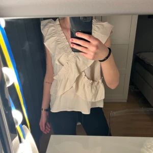 Beige blus från Shein - Säljer en söt beige blus från Shein med volangdetaljer och v-ringning. Blusen är ärmlös och har en luftig passform, perfekt för varmare dagar. Den har en romantisk och feminin stil som passar till många olika tillfällen. Den är aldrig använd💞