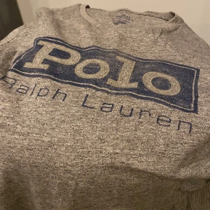 Grå t-shirt från Ralph Lauren stl S  - Säljer en grå t-shirt från Ralph Lauren med stort blått Polo-tryck på framsidan. T-shirten är kortärmad och tillverkad i bomull, perfekt för en avslappnad stil. Passar bra till jeans eller shorts!