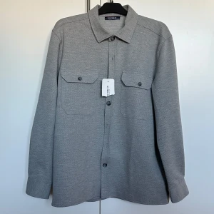Grå overshirt från Southblue - Snygg grå overshirt från Southblue med klassisk krage och knäppning framtill. Två bröstfickor med knappar ger en stilren look. Perfekt för lager-på-lager-stil. Helt ny aldrig använd.