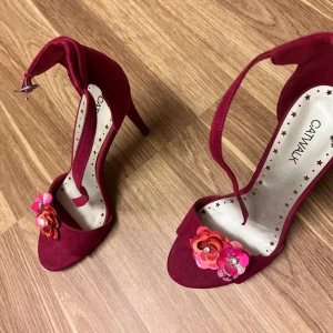 Röda klackskor med blommor från Catwalk - Snygga röda klackskor från Catwalk med öppen tå och dekorativa blommor framtill. Skorna har en elegant rem runt ankeln och en smal klack som ger en stilren look.