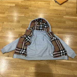 Grå hoodie från Burberry - Säljer en stilren grå hoodie från Burberry med dragkedja och klassiskt rutigt mönster i huvan. Perfekt för en avslappnad och trendig look. Hoodien har långa ärmar och ribbade muddar vid ärmslut och nederkant.
