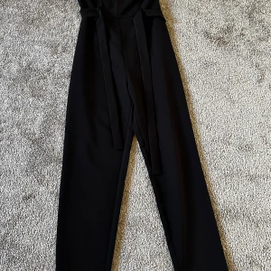 Svart byxdress med knytband - Elegant svart byxdress med smala axelband och knytband i midjan. Den har en stilren design med dragkedja bak och en v-ringad överdel. Perfekt för en chic look.