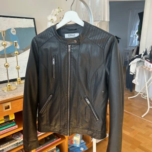 Svart skinnjacka - Snygg svart skinnjacka (fake) med dragkedja och quiltade detaljer på axlarna. Jackan har en stilren design med två framfickor med dragkedja och en knapp vid halsen. Perfekt för en cool look! Den har släppt tyg därav lågt pris.