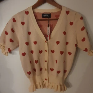 Beige kofta med röda hjärtan från Cider - Säljer en söt beige kofta från Cider med röda hjärtan. Koftan har korta ärmar och volangdetaljer vid ärmslut och nederkant.