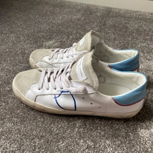 Vita sneakers med blå detaljer från Philippe Model - Snygga vita sneakers från Philippe Model med blå häl och grå mockadetaljer. Skorna har en unik blå design på sidan och klassisk snörning. Perfekta för en stilren look.