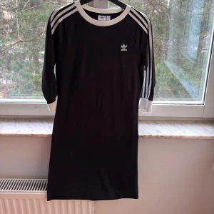 Svart klänning från Adidas - Säljer en svart klänning från Adidas med de klassiska tre ränderna på ärmarna. Klänningen har trekvartsärm och en rund halsringning med vit kant. Perfekt för en sportig look!