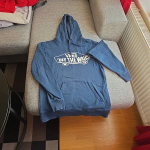 Blå hoodie från Vans - Säljer en blå hoodie från Vans med det klassiska 'Off The Wall' trycket på framsidan. Tröjan har en stor ficka framtill och justerbar huva med dragsko. Perfekt för en avslappnad stil.