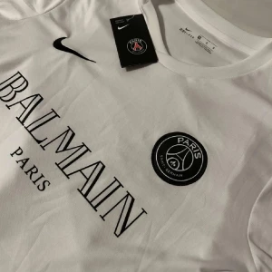 Vit t-shirt från Balmain x PSG - Snygg vit t-shirt från Balmain i samarbete med PSG. Den har en klassisk rund hals och korta ärmar. På framsidan finns Balmain Paris-loggan och PSG-emblemet. Perfekt för dig som älskar mode och fotboll!