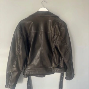 Svart skinnjacka från Zara - Snygg svart skinnjacka från Zara med dragkedja och bälte. Jackan har en klassisk bikerstil med asymmetrisk dragkedja och långa ärmar. Perfekt för en cool och stilren look.