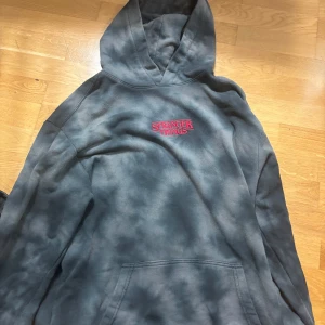 Grå hoodie med Stranger Things-tryck. Lite längre i modellen. - Cool grå hoodie med Stranger Things-tryck i rött och blått. Framsidan har en liten logga medan baksidan har ett stort tryck med karaktärer från serien. Perfekt för fans av serien!