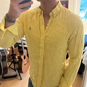 Gul långärmad skjorta från Ralph Lauren - Säljer en 100% linen snygg gul skjorta från Ralph Lauren med klassisk krage och knappar framtill. Skjortan har en broderad logga i blått på bröstet och är tillverkad i ett lätt och bekvämt material.