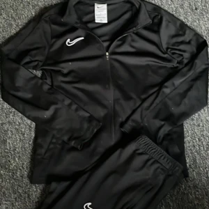 Svart nike tracksuit - Säljer ett svart träningsset från Nike med Dri-FIT-teknologi. Köpte för 950. Setet inkluderar en långärmad tröja med dragkedja och matchande byxor. Perfekt för träning och sportaktiviteter. Nike-loggan är broderad på både tröjan och byxorna. Bra skick inte sönder nånstans
