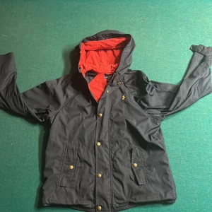Ralph lauren windbreaker - Snygg svart vindjacka från Ralph Lauren med röd insida och huva. Jackan har dragkedja och knappar framtill samt två fickor med knappar. Perfekt för blåsiga dagar!