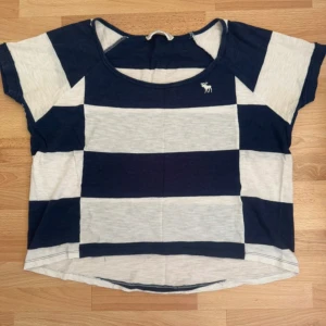 Randig topp från Abercrombie & Fitch XS - Snygg topp från Abercrombie & Fitch i marinblått och vitt med breda horisontella ränder. Modellen är loose fit med korta ärmar och rund halsringning. Liten logga med älg på bröstet. Perfekt för en chill och avslappnad stil.