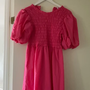 Rosa långklänning med puffärmar - Superfin långklänning i stark rosa med puffiga ärmar och smockad överdel. Klänningen har volangdetaljer och en luftig, flowy passform som känns både trendig och bekväm. Perfekt för dig som vill sticka ut med färg och volym! Inte använts en enda gång den är helt ny, superfin att ta bilder med den, passar super bra att ha på sig den vid en beach