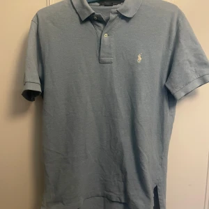 Ljusblå pikétröja från Ralph Lauren - Klassisk ljusblå pikétröja från Ralph Lauren med korta ärmar och vit broderad logga på bröstet. Tröjan har krage, knappar vid halsen och är gjord i mjuk bomull. Perfekt för en clean och stilren look.