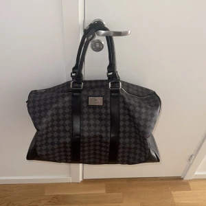 Rutig weekendbag  - Säljer en stor och rymlig weekendbag  i svart och grått rutigt mönster. Väskan har svarta skinnhandtag, detaljer i silver och flera dragkedjor. Perfekt för resor och har en lyxig känsla med metallogo framtill.