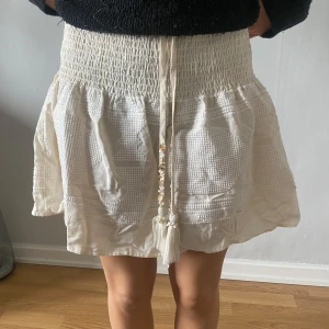 Kortkjol med snörning och tofsar - Supersöt vit kortkjol med smock i midjan och dekorativ snörning med pärlor och tofsar framtill. Kjolen har en strukturerad yta med diskreta ränder och är lätt och luftig, perfekt för varma dagar.