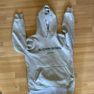 Grå hoodie från Björn Borg - Snygg ljusgrå hoodie från Björn Borg med svart logga framtill. Klassisk modell med huva, känguruficka och ribbade muddar. Perfekt för chill dagar eller när du vill ha en sportig vibe. Tillverkad i mjuk bomullsblandning för extra komfort. Kan förekomma katt hår för finns katt i huset
