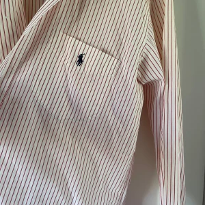 Randig skjorta från Polo Ralph Lauren - Vit skjorta med röda smala ränder från Polo Ralph Lauren. Klassisk krage, långärmad och med knappar framtill. Bröstficka med broderad logga i mörkblått. Skjortan är i bomull och har en tidlös, clean look.