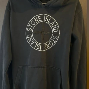 Svart Stone Island hoodie XXL - Svart hoodie från Stone Island i storlek XXL med stort broderat logotyptryck på bröstet. Klassisk huva, känguruficka och mjukt material som känns skönt mot huden. Perfekt för dig som gillar streetwear och vill sticka ut med en ikonisk look.