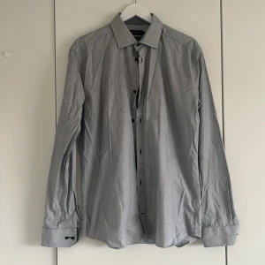 Grå smalrandig skjorta från Riley - Snygg vit skjorta från Riley i slim fit med smala gråa ränder och svarta knappar. Klassisk krage och långa ärmar med breda manschetter. Skjortan är gjord i bomull och har diskret mönstrat tyg på insidan av kragen.