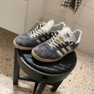 Adidas Gazelle i mörkblå mocka - Snygga Adidas Gazelle sneakers i mörkblå mocka med vita detaljer och klassiska tre ränder på sidan. Skorna har vit plös och snörning samt gummisula med mönster undertill. 
