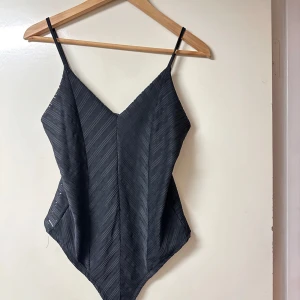 Svart randig bodysuit med tunna band - Säljer en svart bodysuit med tunna axelband och v-ringad hals. Tyget har ett diskret randigt mönster med genomskinliga detaljer som ger en cool effekt. Perfekt att styla med jeans eller kjol för en trendig look.