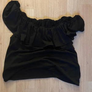 Svart offshoulder topp med volang - Snygg svart offshoulder topp med bred volang runt hela överdelen. Perfekt för dig som gillar en trendig och feminin stil. Toppen är ärmlös och har en elastisk kant upptill som håller den på plats.