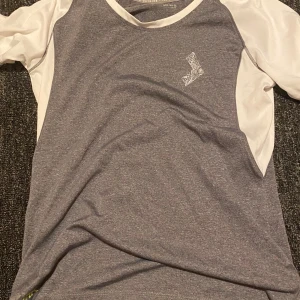 Grå och vit sportig t-shirt - Sportig t-shirt från lager i grått med vita ärmar och sidor. T-shirten har korta ärmar och en liten vit logga på bröstet. Perfekt för träning eller aktiva dagar. Materialet känns lätt och andas bra.