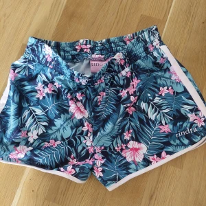 Blommiga shorts från Tindra - Snygga shorts från Tindra med tropiskt blommönster i blått, rosa och vitt. De har elastisk midja och vita detaljer längs sidorna. Perfekta för sommaren och strandhäng. Lätta och bekväma med en loose passform.