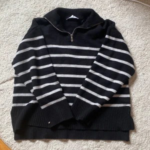 Randig svart stickad tröja från Zara - Svart och grå randig stickad tröja från Zara med halv dragkedja i halsen. Tröjan har ribbade muddar och hög krage, perfekt för kyliga dagar. Långärmad och i mjukt material som känns skönt mot huden.