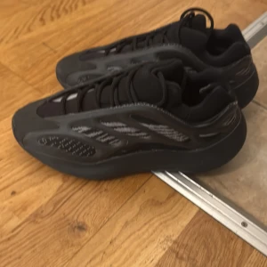 Svarta Adidas Yeezy 700 V3 sneakers - Säljer ett par svarta Adidas Yeezy 700 V3 sneakers med futuristisk design och reflekterande detaljer. Skorna har snörning, rund tå och en chunky sula. Ovandelen är i syntet och mesh med grå och svarta inslag. Perfekta för dig som gillar streetwear och vill sticka ut.