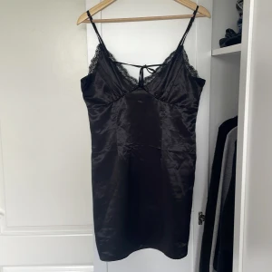Svart satin klänning från H&M  - Svart klänning från H&M i glansigt satinliknande material. Klänningen har tunna axelband, spetsdetaljer vid urringningen och en liten knytning bak. Hör av er vid frågor!💗